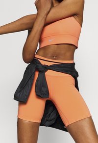 Sujetador deportivo naranja y pantalones cortos a juego con una chaqueta ligera negra atada a la cintura. El sujetador tiene un pequeño logo de Nike.
