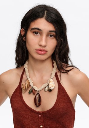 Mujer con cabello largo y oscuro que lleva una blusa marrón sin mangas y un collar grueso hecho de cuerda trenzada y grandes conchas marinas.