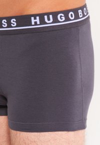 Dunkelgraue Boxershorts aus einem weichen, elastischen Stoff. Mit einem weißen Gummibund, auf dem "HUGO BOSS" steht. Steppnähte sichtbar.