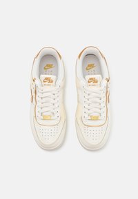 Les baskets Nike Air Force 1 présentent une tige en cuir crème, des accents dorés, un bout perforé et une semelle en caoutchouc blanche. Lacets et logo visibles.