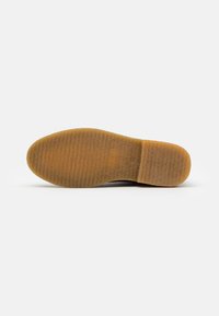 Vue de la semelle de chaussure montrant un matériau en caoutchouc brun avec un motif texturé antidérapant, un talon plat et un bout arrondi sur fond neutre.