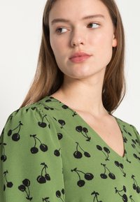 Blusa verde com decote em V, apresentando um padrão de cerejas pretas e mangas bufantes. Feita de tecido leve com uma textura suave.