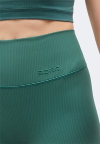 Κοντινό πλάνο ενός ατόμου που φοράει πράσινα ριμπ πλεκτά κολάν ψηλόμεσα με το λογότυπο "BORG" σε ανάγλυφη μορφή στη μέση.