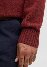 Pull rouge texturé avec des poignets côtelés, porté sur une main tatouée. Le tissu semble doux, avec une coupe décontractée, superposé sur un pantalon marine.