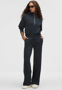Ciemna, dopasowana bluza z suwakiem do połowy i kapturem, w zestawie z dopasowanymi szerokimi spodniami dresowymi. Gładki materiał, przednie kieszenie i swobodny design.