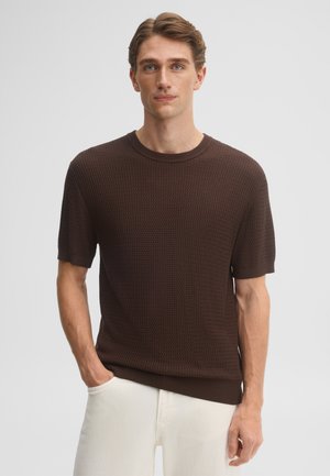 Junger Mann mit heller Haut und braunen Haaren trägt einen dunkelbraunen, strukturierten Kurzarmpullover und weiße Hose und steht vor einem schlichten Hintergrund.