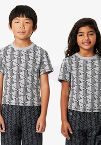 Due bambini indossano t-shirt grigie con un motivo geometrico nero, dotate di maniche corte e scollo rotondo. Entrambi indossano pantaloni neri a fantasia.