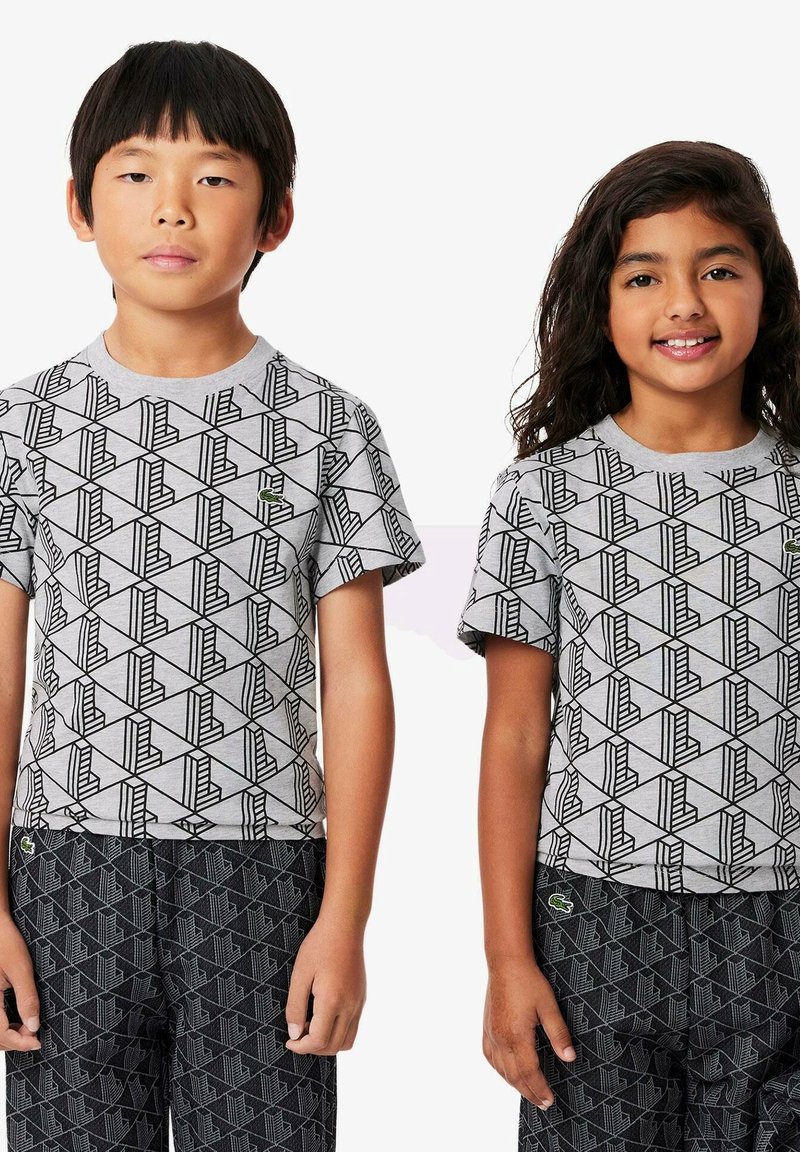 Due bambini indossano t-shirt grigie con un motivo geometrico nero, dotate di maniche corte e scollo rotondo. Entrambi indossano pantaloni neri a fantasia.