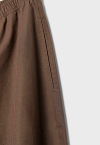 Pantalon en tissu marron avec poche fendue latérale et taille élastique, présenté sur un fond gris clair.