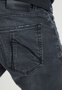 Jean gris foncé en denim avec une finition texturée. Comprend une poche arrière visible avec des coutures contrastantes et une étiquette de logo.
