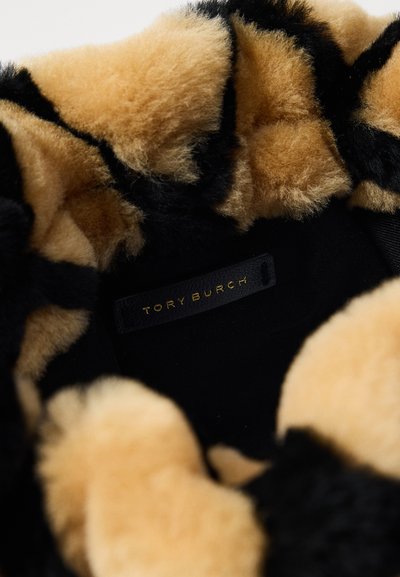 Melnas un beiges mākslīgās kažokādas interjers ar mīkstu, teksturētu sajūtu. Redzama melna etiķete ar "TORY BURCH" zeltītā rakstībā.