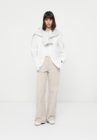 Donna con capelli scuri, che indossa una camicia bianca e pantaloni beige, con un maglione grigio annodato attorno alle spalle, in piedi contro uno sfondo bianco semplice.