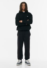 Svart huvtröja med ficka framtill och logotyp, kombinerad med svarta sweatpants. Skorna har en grå och vit design med snörning. Enkelt, avslappnat utseende.