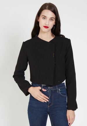 Chaqueta negra corta con mangas largas, con un cuello en ángulo y cierre de botones, hecha de tela suave. Llevada con jeans de mezclilla oscuros.