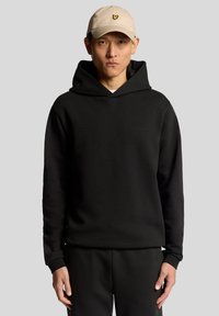 Felpa nera con cappuccio a coulisse, texture morbida e un piccolo logo sul petto. Abbinata a pantaloni neri semplici e un berretto beige.