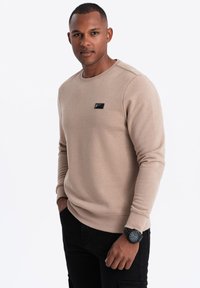 Felpa beige con colletto rotondo, polsini a coste e una toppa nera sul petto, abbinata a pantaloni neri e un orologio da polso nero.
