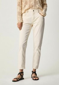 Jeans blancs à coupe droite et poches avant, associés à un pull en maille beige ajourée et des sandales à lanières noires.