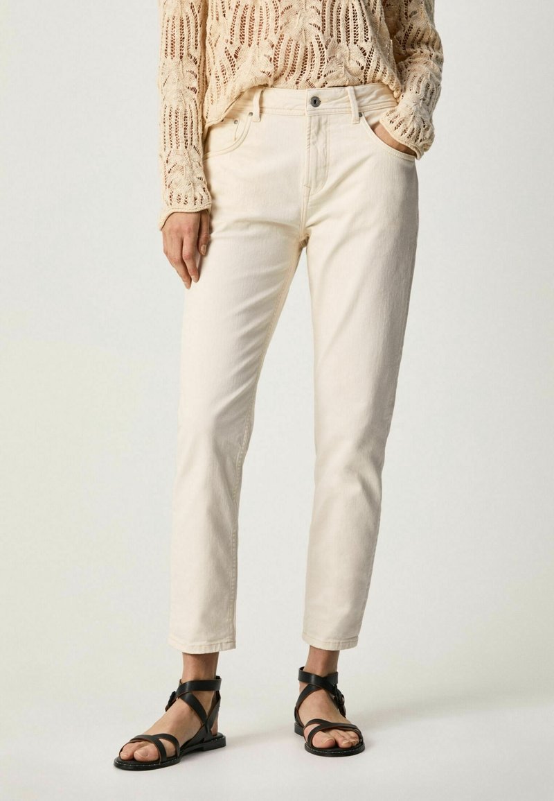 Jeans blancs à coupe droite et poches avant, associés à un pull en maille beige ajourée et des sandales à lanières noires.