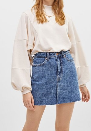 Kobieta ubranawu kremową, oversize'ową bluzę z bufiastymi rękawami oraz wysokim stanem mini spódnicę z niebieskiego denimu na jednolitym tle.