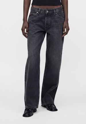 Personne portant un jean noir délavé et ample et des chaussures noires à bout carré, debout devant un fond clair uni.