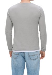 Homme portant une chemise à manches longues grise claire texturée sur un débardeur blanc et un jean en denim bleu clair, vu de dos.