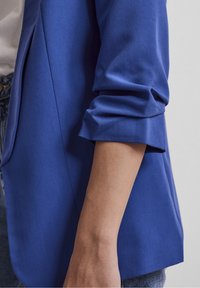 Blazer bleu avec une texture lisse et mate ; manches retroussées ; une poche latérale ; coupe cintrée ; revers nets ; associé à un jean clair.