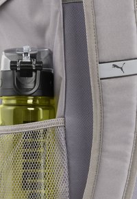 Grijze rugzak met een mesh zijvak dat een heldere en zwarte plastic waterfles vasthoudt, die een gele basis en een veilige deksel heeft.