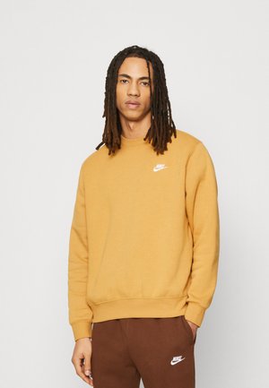 Sudadera de Nike en amarillo mostaza, con un cuello redondo, mangas largas y un pequeño logo blanco en el pecho. Textura suave, ajuste relajado.