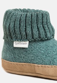 Pantofola verde morbida con bordo a coste, parte superiore in tessuto a effetto lana, suola in suede marrone chiaro e piccolo cartellino bianco etichettato "Gottstein" sul lato.