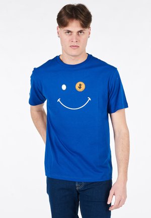 T-shirt in cotone blu con una grande grafica di un volto sorridente bianco e un logo arancione rotondo sull'area sinistra del petto. Maniche corte, design a girocollo.