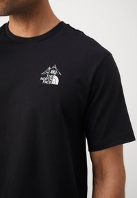 Black cotton t-shirt featuring a small, embroidered mountain and logo design in white and teal on the left chest area. Short sleeves.