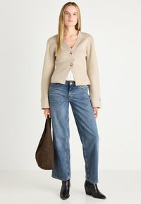 Kvinne iført beige knapped cardigan, hvit topp, blå vide jeans, svarte spisse støvler, som holder en stor mørkebrun veske, står foran en hvit bakgrunn.
