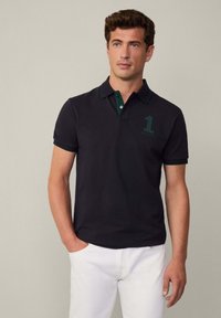 Čierne polo tričko so zeleným golierom a detailmi na gombíkoch, na hrudi s veľkým zeleným logom "1". Nosí sa s bielymi nohavicami.
