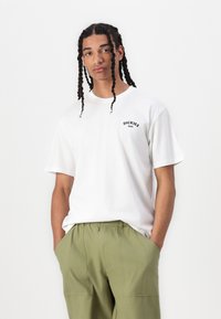 Fehér pamut póló, bal oldalon kicsi fekete "Dickies" logóval, olíva zöld, elasztikus derékú nadrággal párosítva.