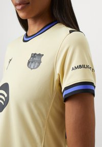 Camiseta de fútbol beige con un diseño texturizado, que presenta un escudo negro del Barcelona, el logo de Spotify y acentos en rayas azul y negro en las mangas.