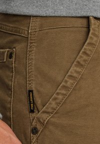 Pantalones de tela marrón con textura, costuras visibles, una pequeña etiqueta negra y amarilla "PME Legend" y un detalle de bolsillo con botón.