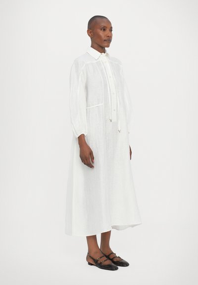 Abito lungo bianco in lino con colletto, bottoni frontali e due tasche laterali. Accentuato da un dettaglio con fiocco e maniche larghe a sbuffo.