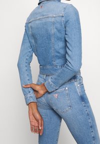 Světlá modrá riflová bunda s přiléhavým střihem, dlouhými rukávy a límečkem. Má logo nášivku a prošité detaily na zádech a manžetách.