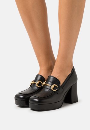 Escarpins en cuir noir à plateforme avec détail en chaîne dorée et boucle circulaire, portés sur des pieds sur un fond uni.