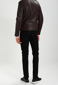 Bruine leren motorjack, aangesloten zwarte jeans en zwarte sneakers. Het jack heeft schouderepauletten; de jeans hebben achterzakken.