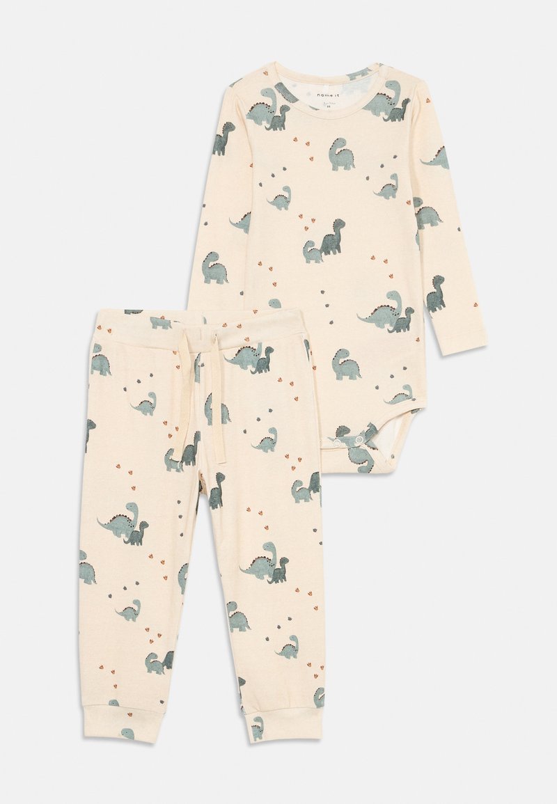 Cream baby pajamas with long-sleeve bodysuit and pants, featuring blue and gray dinosaur prints and small footprints.