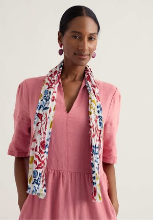 Ženska s temnimi lasmi, povezanimi nazaj, nosi roza obleko, rdeče uhane in belo ruto z rdečimi, modrimi in rumenimi cvetličnimi vzorci, ogrnjeno okoli vratu.