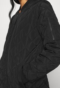 Černá prošívaná bomber bunda s kapsou na zip na hrudi, žebrovanými manžetami a výstřihem a strukturovaným povrchem. Kompaktní a lehký design.