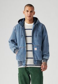Veste en denim avec capuche, de couleur bleue, avec des poignets élastiques et des poches devant, portée sur une chemise rayée. Modèle portant un pantalon en velours côtelé vert.