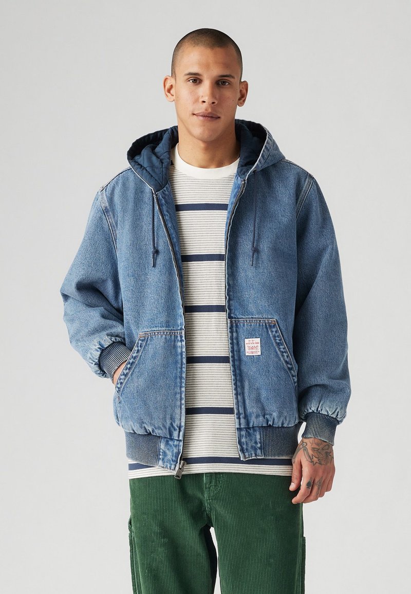 Veste en denim avec capuche, de couleur bleue, avec des poignets élastiques et des poches devant, portée sur une chemise rayée. Modèle portant un pantalon en velours côtelé vert.