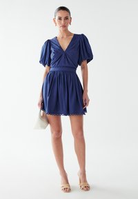 Navyblaues Kleid mit Puffärmeln, V-Ausschnitt und gerafftem Rock. Abgeschlossen mit gewelltem Saum. Modell hält eine weiße Handtasche.