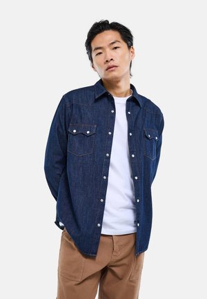 Jeune homme aux cheveux noirs portant une chemise en denim bleu foncé sur un t-shirt blanc et un pantalon marron, debout les mains derrière le dos.