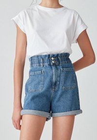 Persona que lleva pantalones cortos de mezclilla azul de talle alto y con puños, con bolsillos frontales y una camiseta blanca de manga corta, frente a un fondo liso.