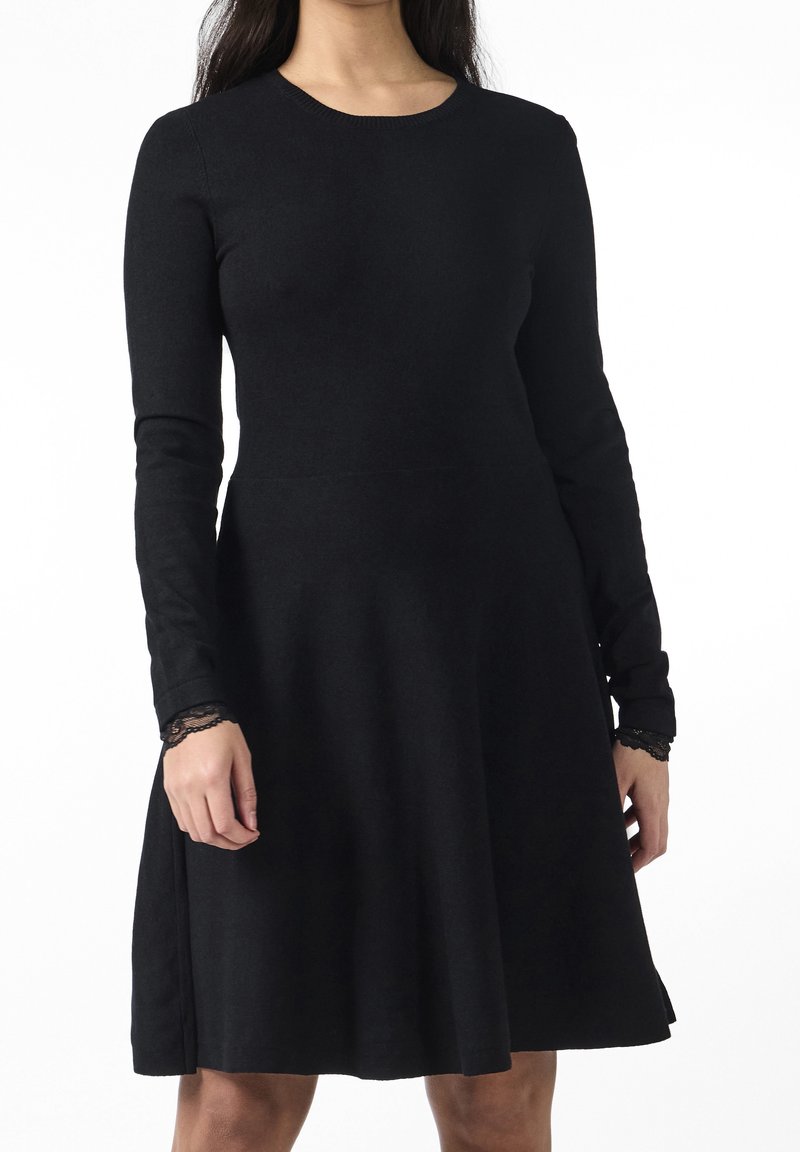 Femme portant une robe noire à manches longues avec une bordure en dentelle aux poignets, debout devant un fond blanc, mains détendues le long du corps.
