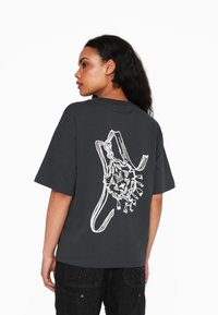 Grå oversized t-shirt lavet af bomuld, med et hvidt grafisk tryk af et løvedesign på ryggen. Korte ærmer og rund halsudskæring.